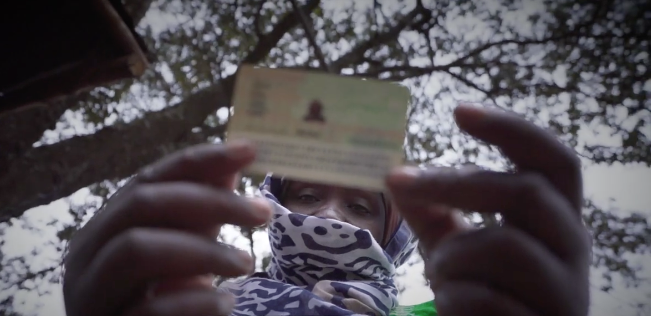 A woman with a headscarf covering her mouth looks down at her ID while standing under a tree.