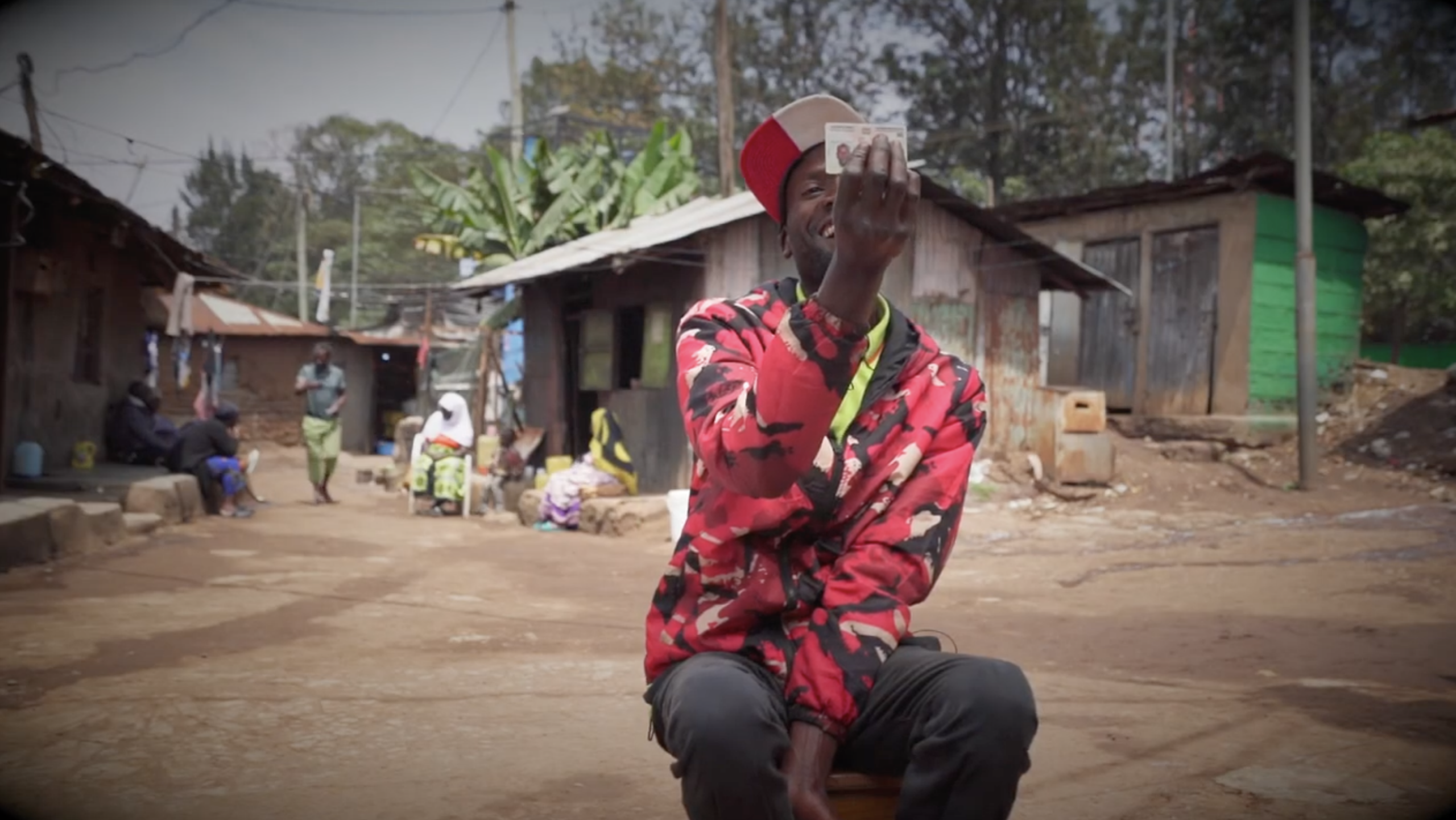A man sits outside in a village smiling up at his ID card.